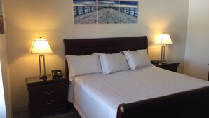 A neatly made bed with white linens, two bedside tables with lamps, a wooden headboard, and a painting of a pier above the bed.
