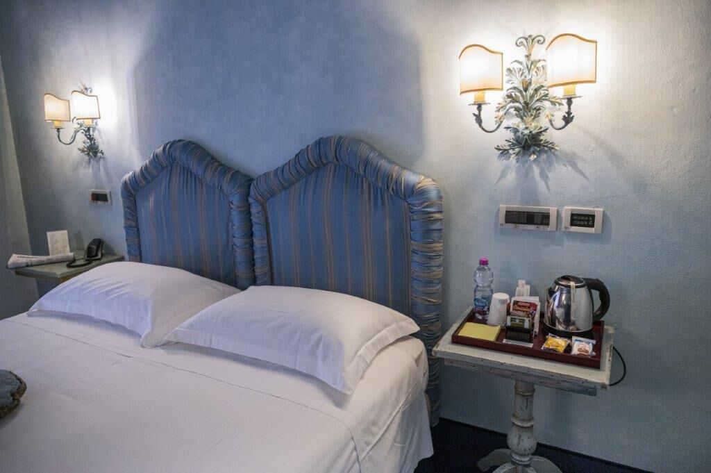Camera d'albergo con un letto grande, due lampade a parete, un comodino con un vassoio di articoli da toilette e un termostato.