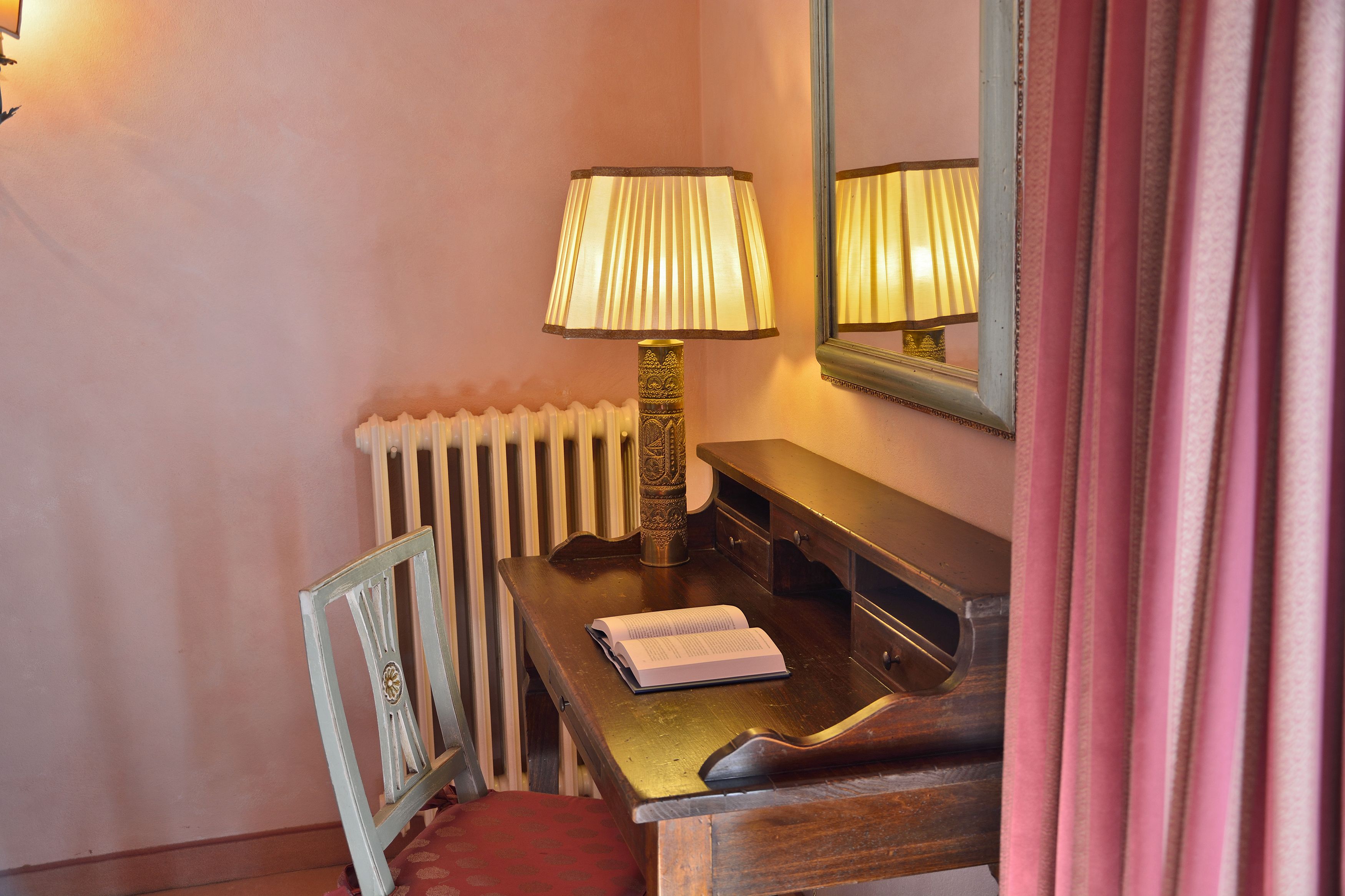 A corner of a room with a wooden desk, a lamp, a radiator, a mirror, and a chair.
