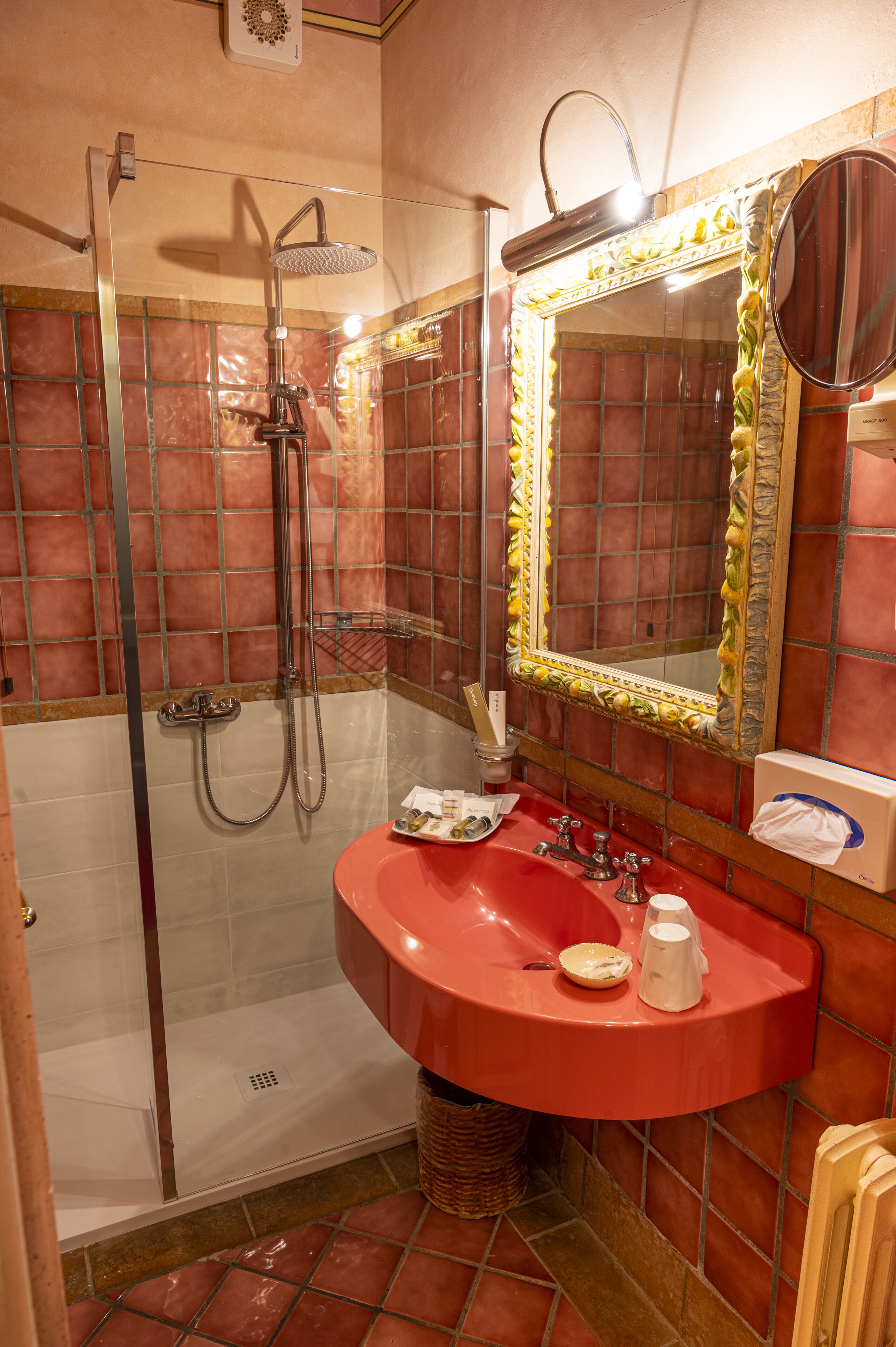 A bathroom with a glass-enclosed shower, a red sink, and a mirror with a decorative frame.