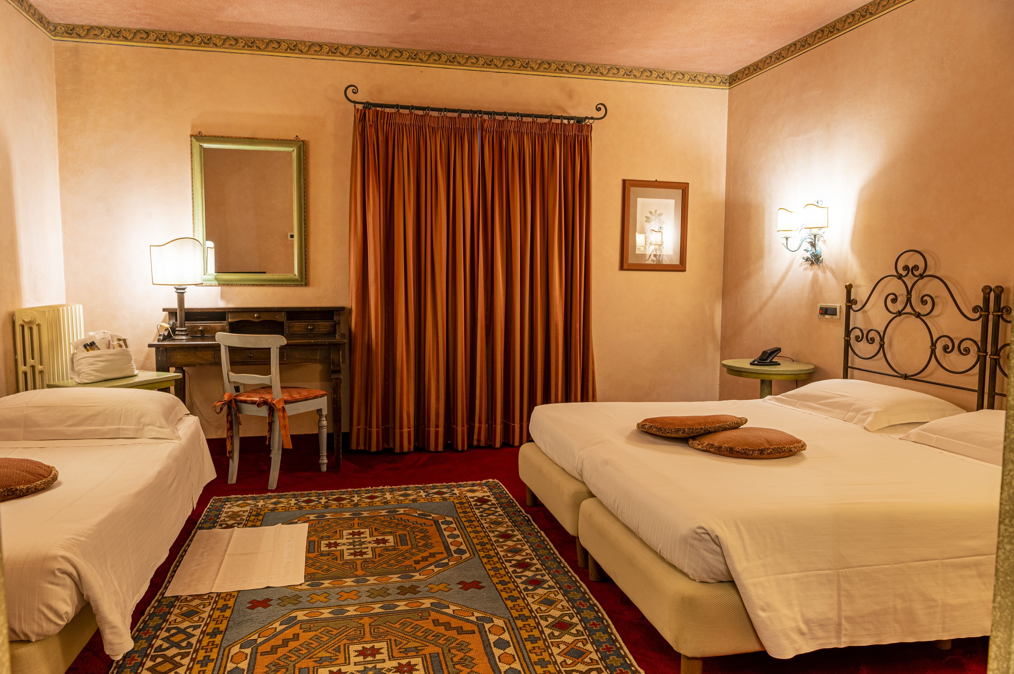 A hotel room with two beds, a desk, a chair, a small table, a lamp, a mirror, and a curtain.