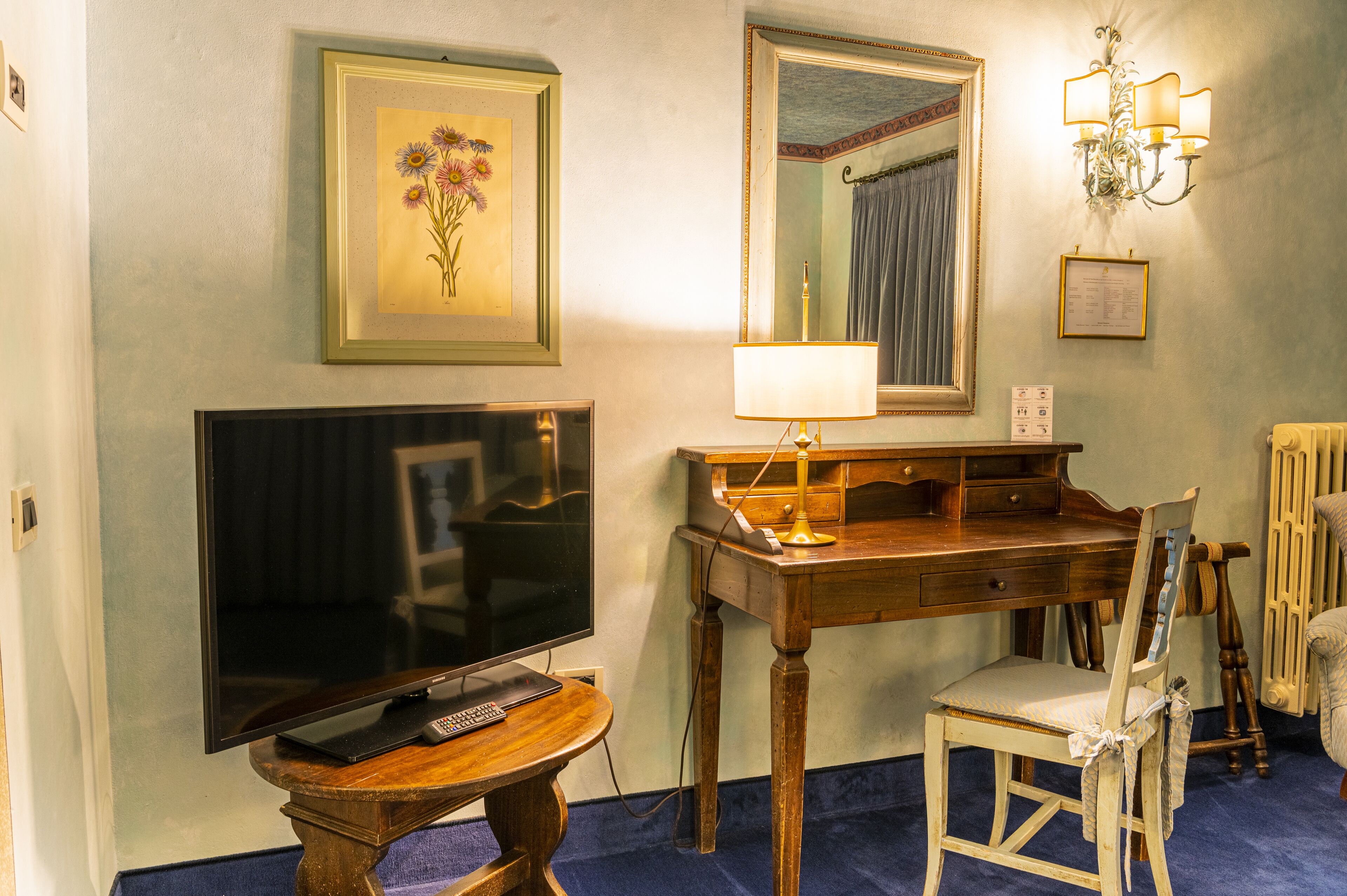Quarto com televisão de tela plana, escrivaninha de madeira com luminária, cadeira, obra de arte floral emoldurada, espelho e lustre.