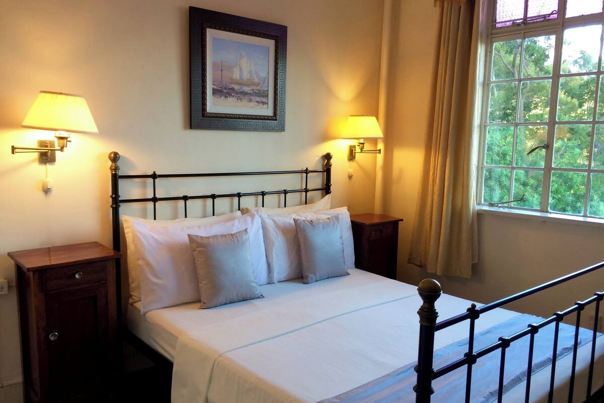 A neatly made bed with white linens, two bedside tables with lamps, a framed picture on the wall, and a window with curtains.