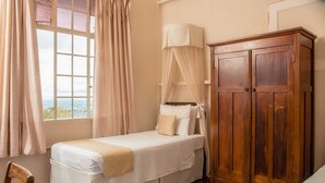 A single bed with a canopy, a wooden wardrobe, and a view through a window with curtains.