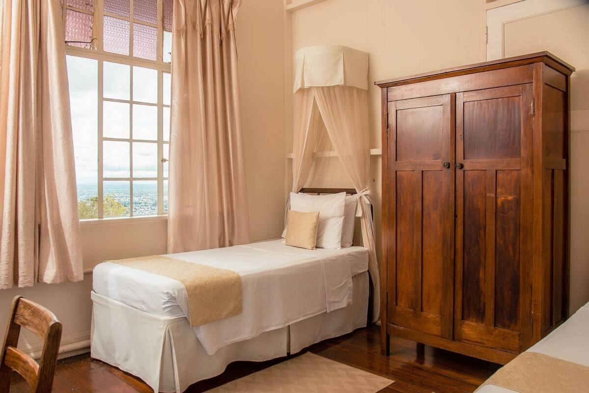 A single bed with a canopy, a wooden wardrobe, and a view through a window with curtains.