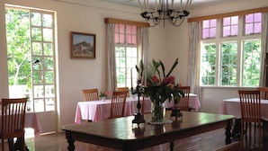 A dining area with a large wooden table, chairs, and a chandelier. There are windows with curtains and a painting on the wall.