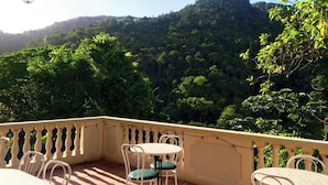 A terrace with a view of a forested hill, white chairs, and a table.
