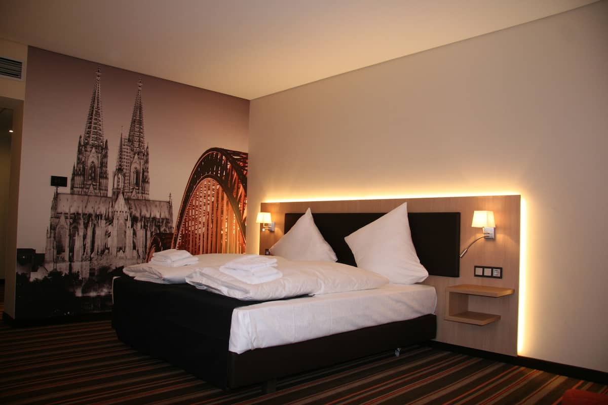 A hotel room with a large bed, a wall-mounted lamp, and a mural of a cathedral.