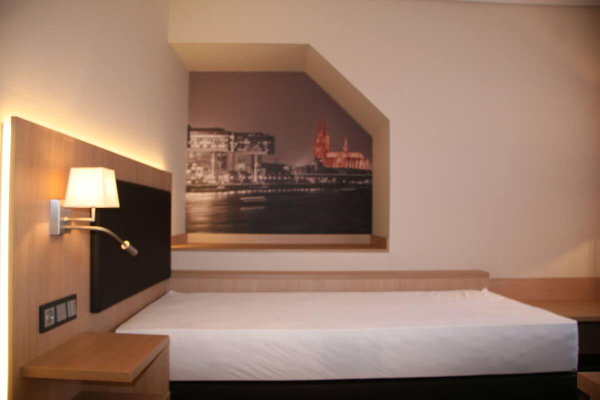 A hotel room with a bed, a nightstand, a wall-mounted lamp, and a cityscape painting.