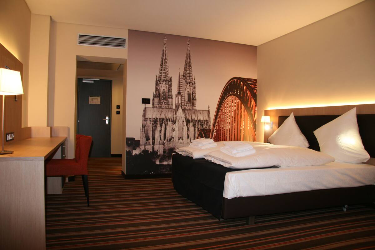 A hotel room with a bed, a desk, a chair, and a large wall mural of a cathedral.