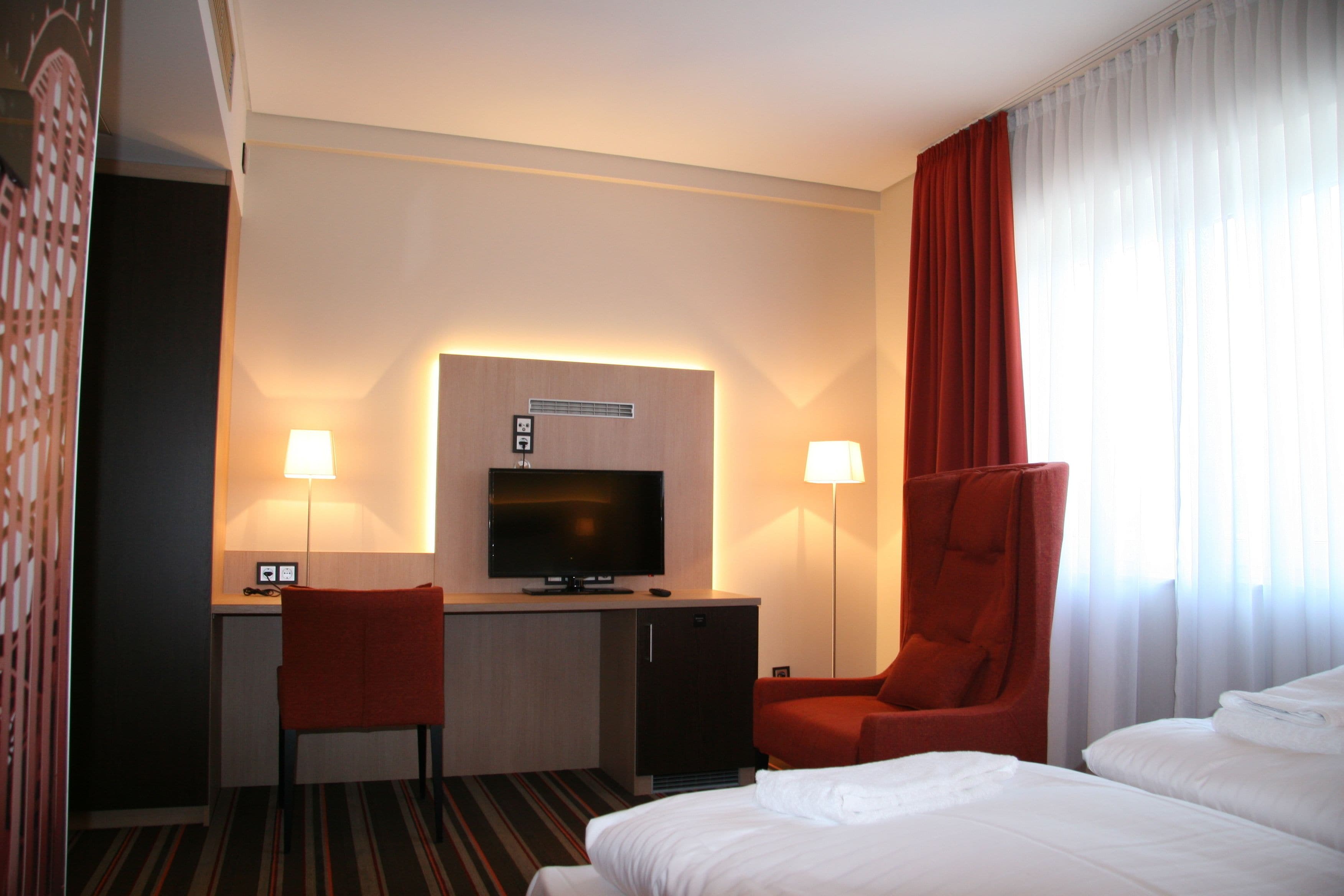 A hotel room with a bed, a desk with a TV, a chair, and a lamp.