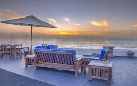 A beachside lounge with wooden furniture and a large umbrella, overlooking the ocean.