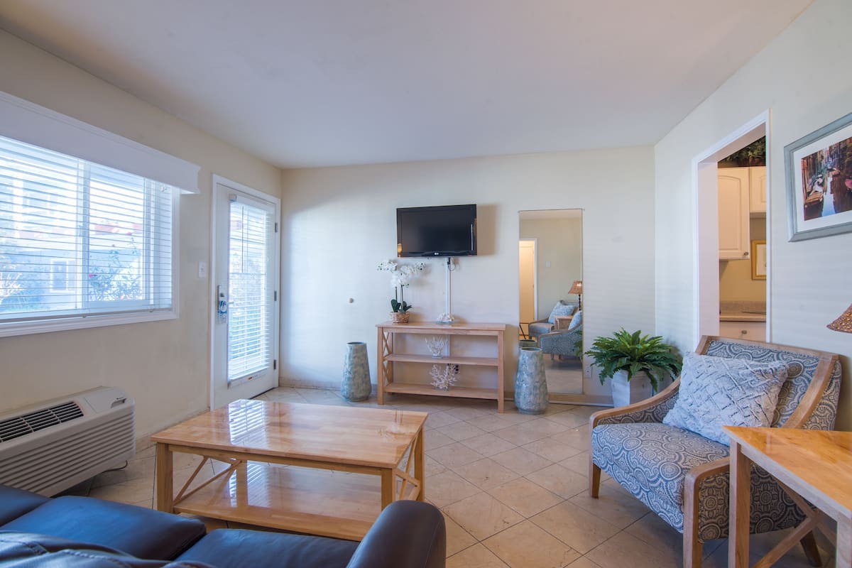 A living room with a sofa, a coffee table, a TV mounted on the wall, a potted plant, and a chair with a cushion.