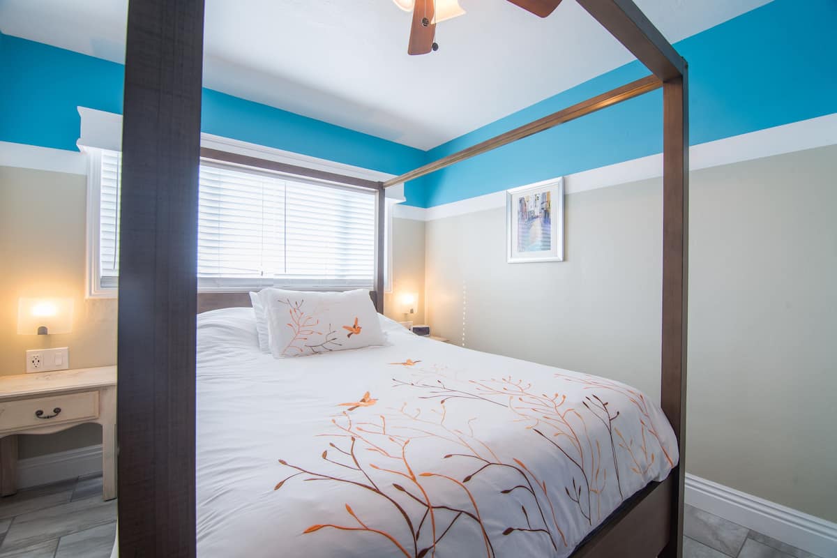 A bedroom with a four-poster bed, a nightstand, a bedside table, and a window with blinds.