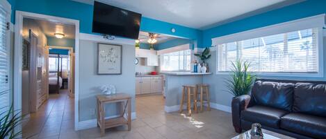A modern living room with a brown leather sofa, a wooden coffee table, and a kitchen area with white cabinets and a blue accent wall.
