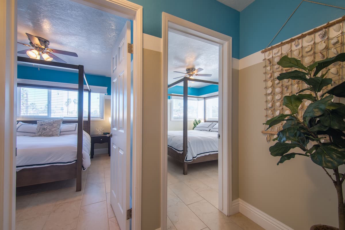 A bedroom with a four-poster bed, a ceiling fan, and a large window with blinds.