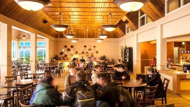 A spacious dining area with wooden tables and chairs, a high ceiling with exposed beams, and a view of the outdoors through large windows.