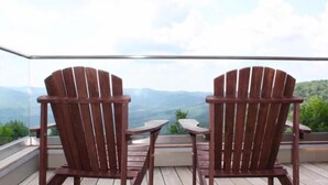 Two wooden Adirondack chairs facing each other on a balcony with a glass railing, overlooking a mountainous landscape.