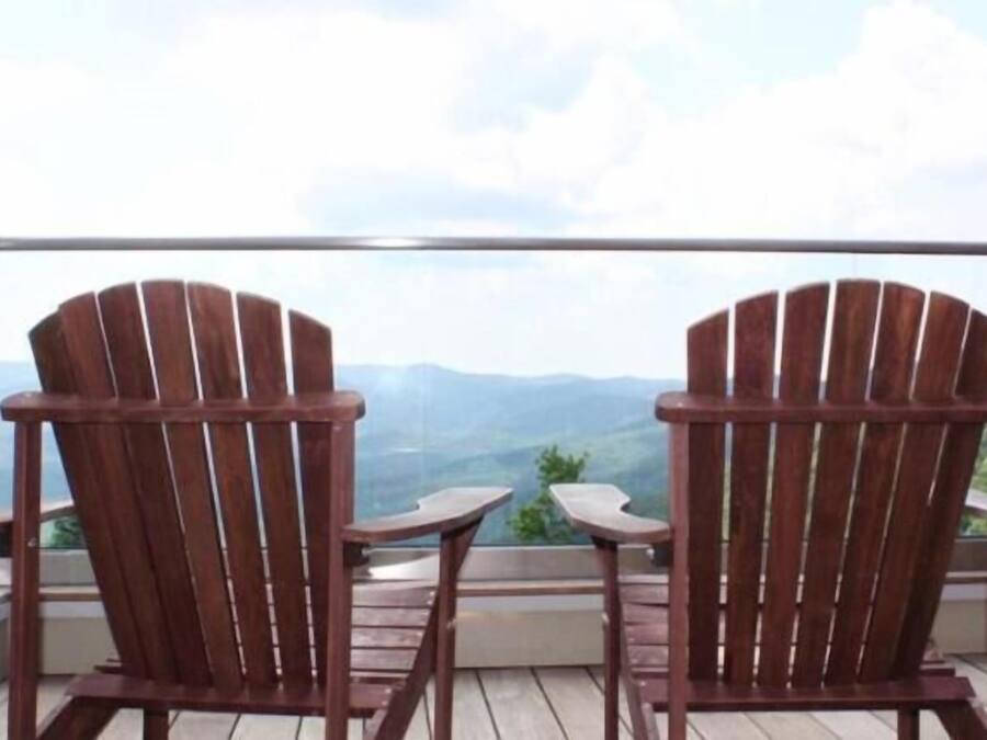 To træstole af Adirondack-design står overfor hinanden på en balkon med gelænder i glas, med udsigt over et bjerrelandskab.