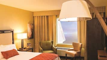 Quarto de hotel com uma cama, uma escrivaninha, uma cadeira, um abajur, uma janela com cortinas e vista para o céu.
