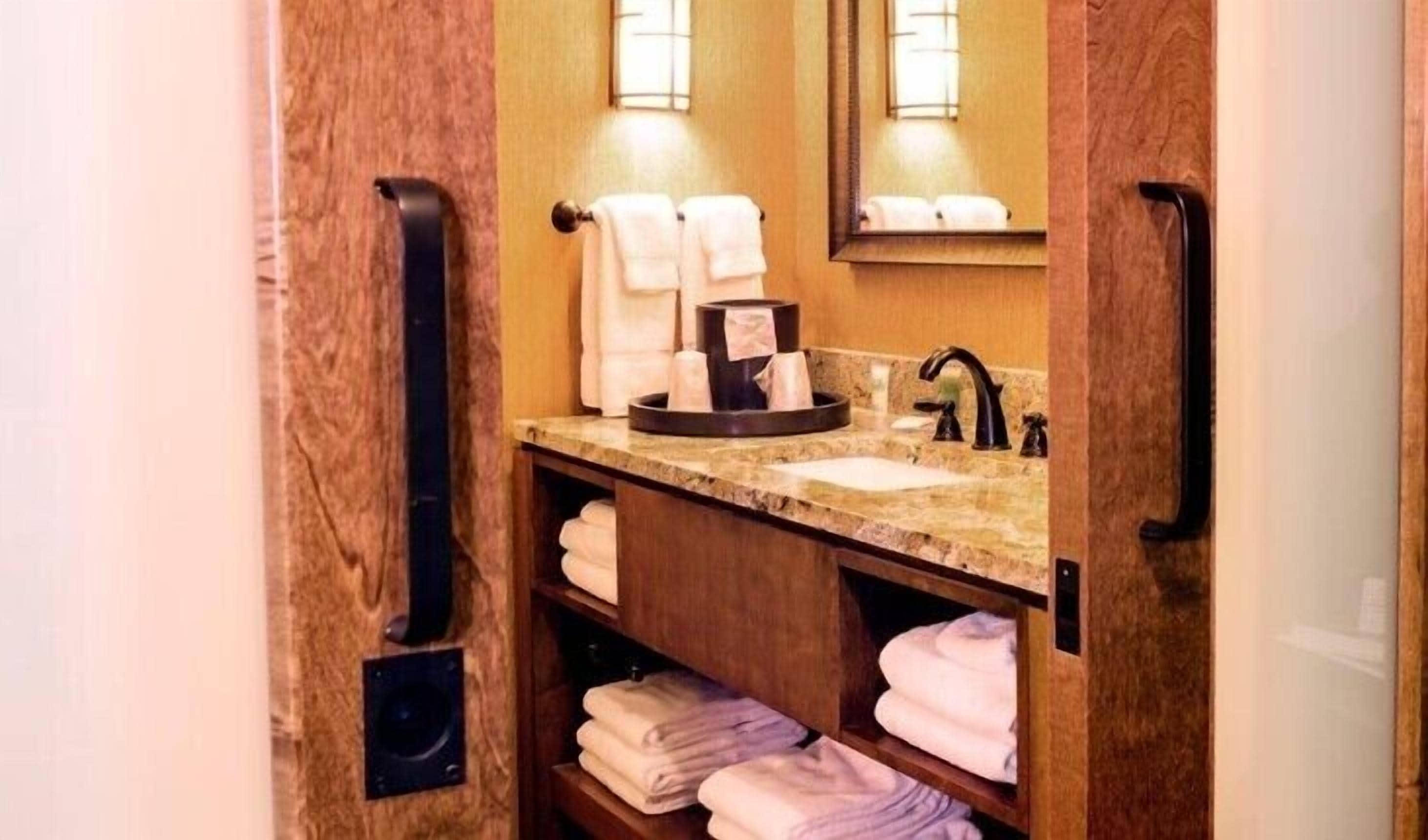 Une petite salle de bain dotée d’un comptoir en marbre, d’un lavabo et d’un miroir. Des serviettes sont soigneusement pliées et rangées sur des étagères, et un porte-serviettes noir est fixé au mur.
