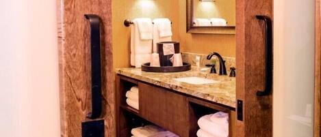 Une petite salle de bain dotée d’un comptoir en marbre, d’un lavabo et d’un miroir. Des serviettes sont soigneusement pliées et rangées sur des étagères, et un porte-serviettes noir est fixé au mur.