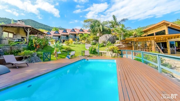 A pool area with wooden decking, lounge chairs, and a view of resort buildings and greenery. - Pousada do Taxo (Garopaba)