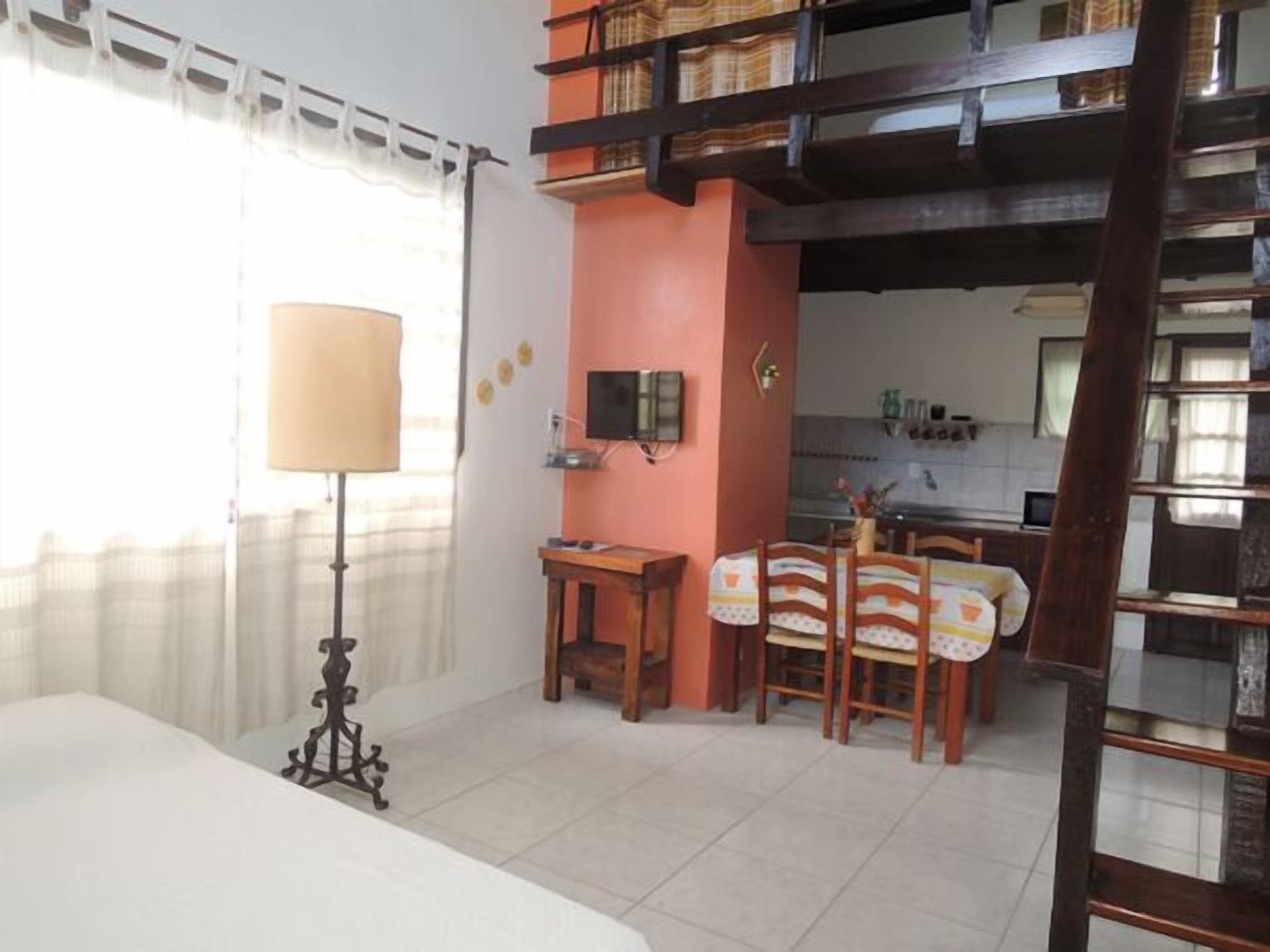 A room with a bed, a desk, a chair, a lamp, and a kitchen area with a table and chairs.