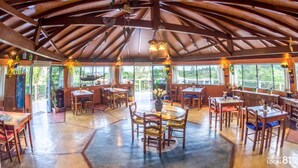 A spacious dining area with multiple tables and chairs, large windows offering views of the outdoors, and a high ceiling with exposed beams. - Pousada do Taxo (Garopaba)