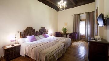 Habitación de hotel con una cama grande, suelo de madera, araña, escritorio con silla y televisor en la pared.