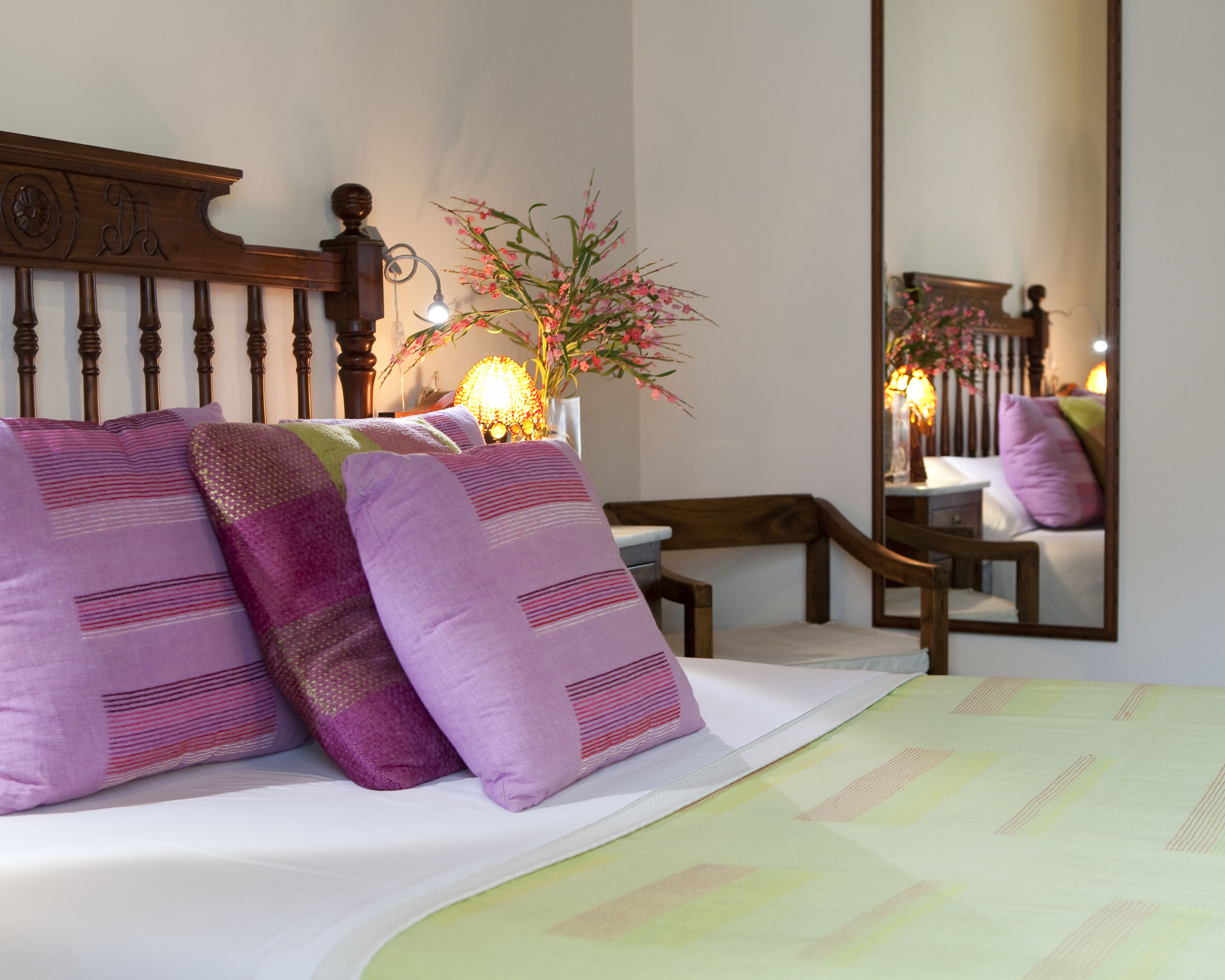 A neatly made bed with purple pillows, a bedside lamp, and a mirror reflecting the room's decor.