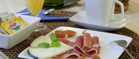 Une table dressée pour le petit-déjeuner avec une assiette de fromage, de jambon de Parme et de tomates, un verre de jus d’orange, une tasse de café et un panier de pain.