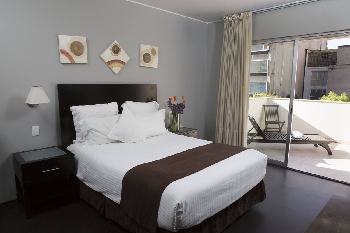 A hotel room with a bed, a nightstand, a lamp, a vase with flowers, and a view of the outside balcony.