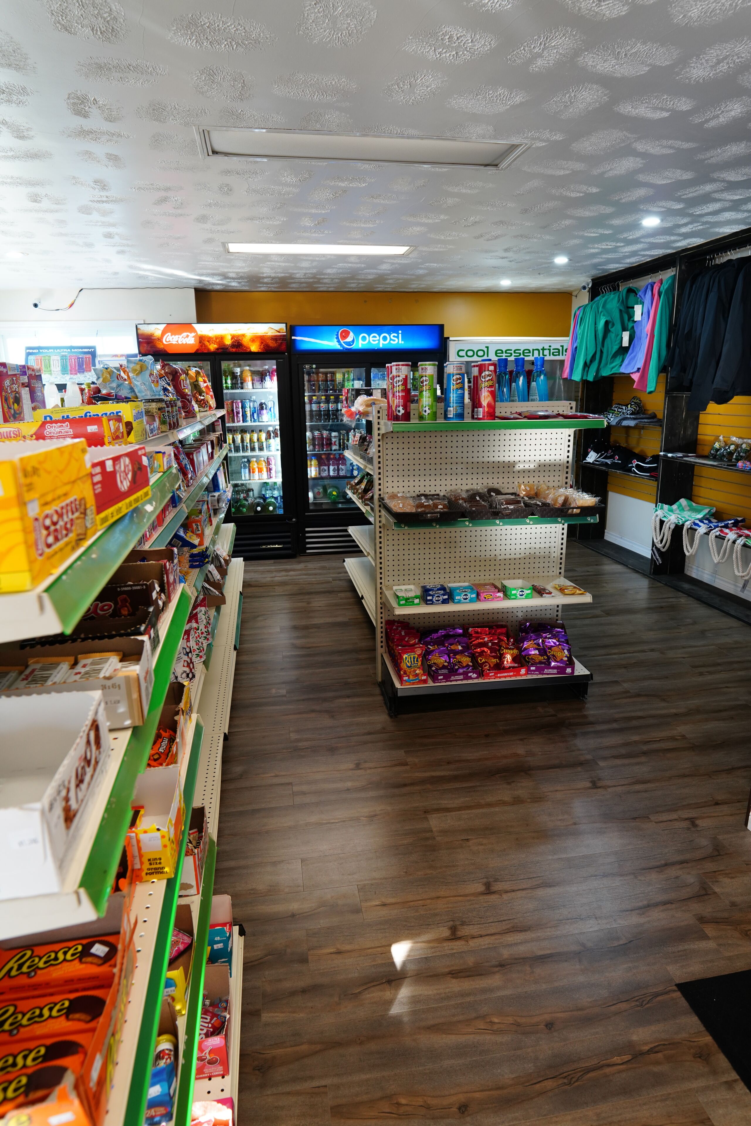 A store with shelves stocked with snacks, a Pepsi refrigerator, and a section with clothing items.