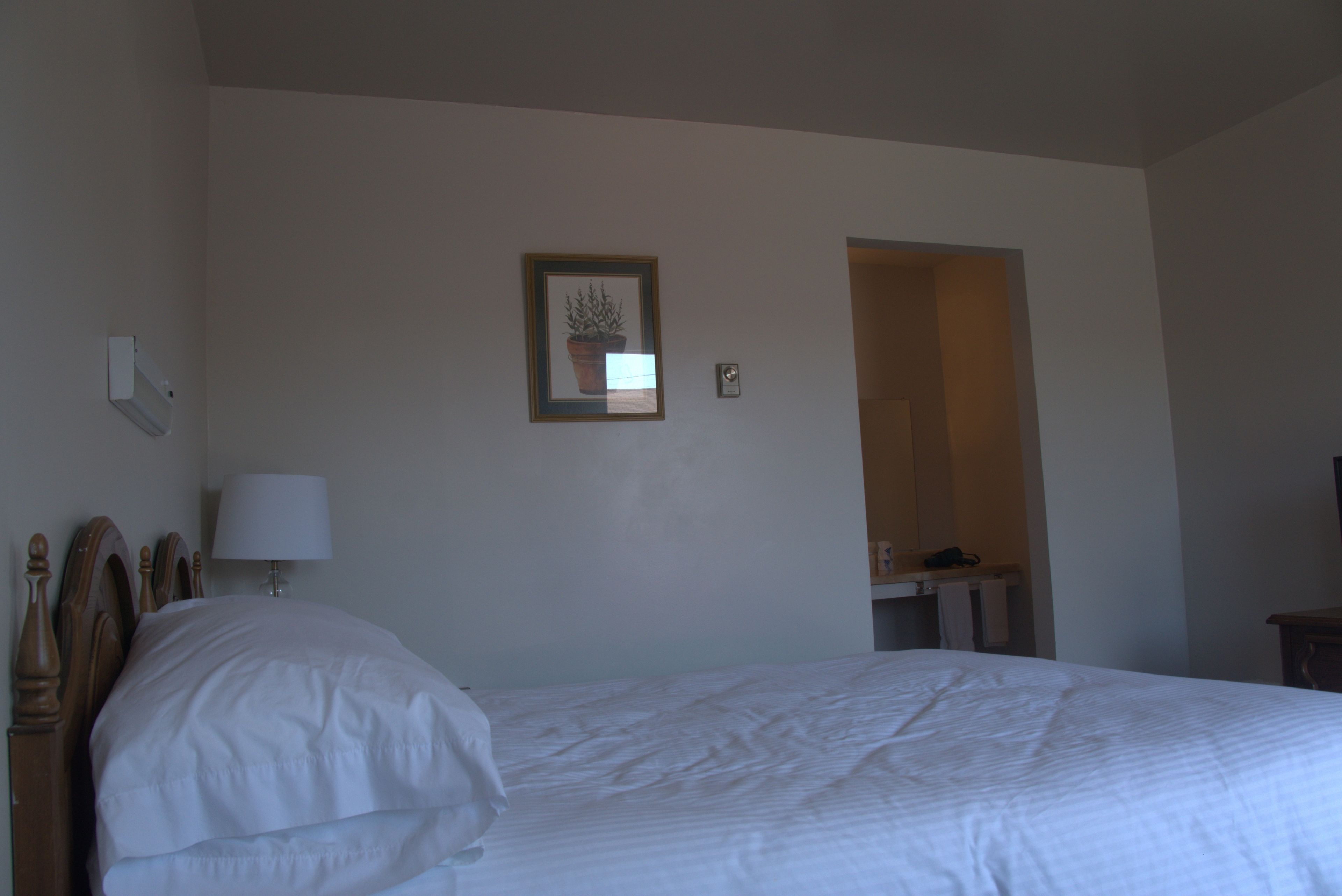 A hotel room with a single bed, a bedside table with a lamp, a framed picture on the wall, and an open doorway to another room.