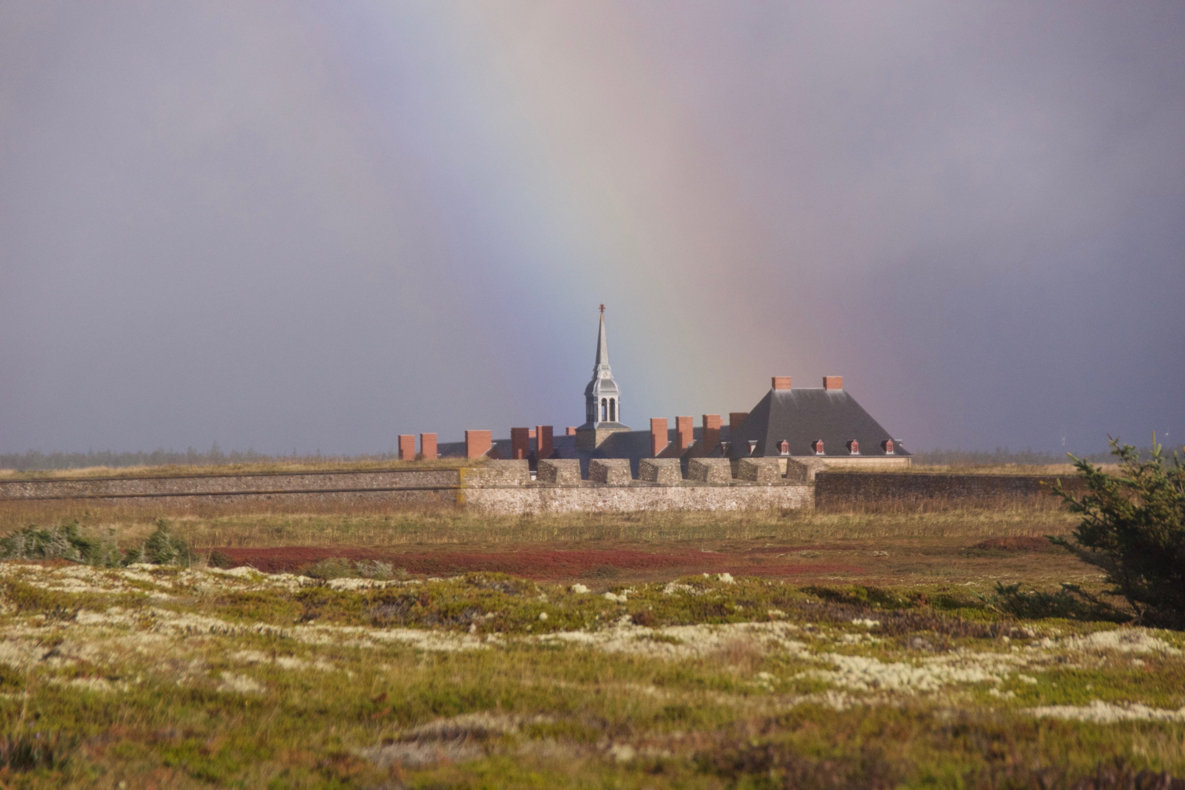 A church with a tall steeple and a bell tower, surrounded by a field with a fence and a small hill in the background.