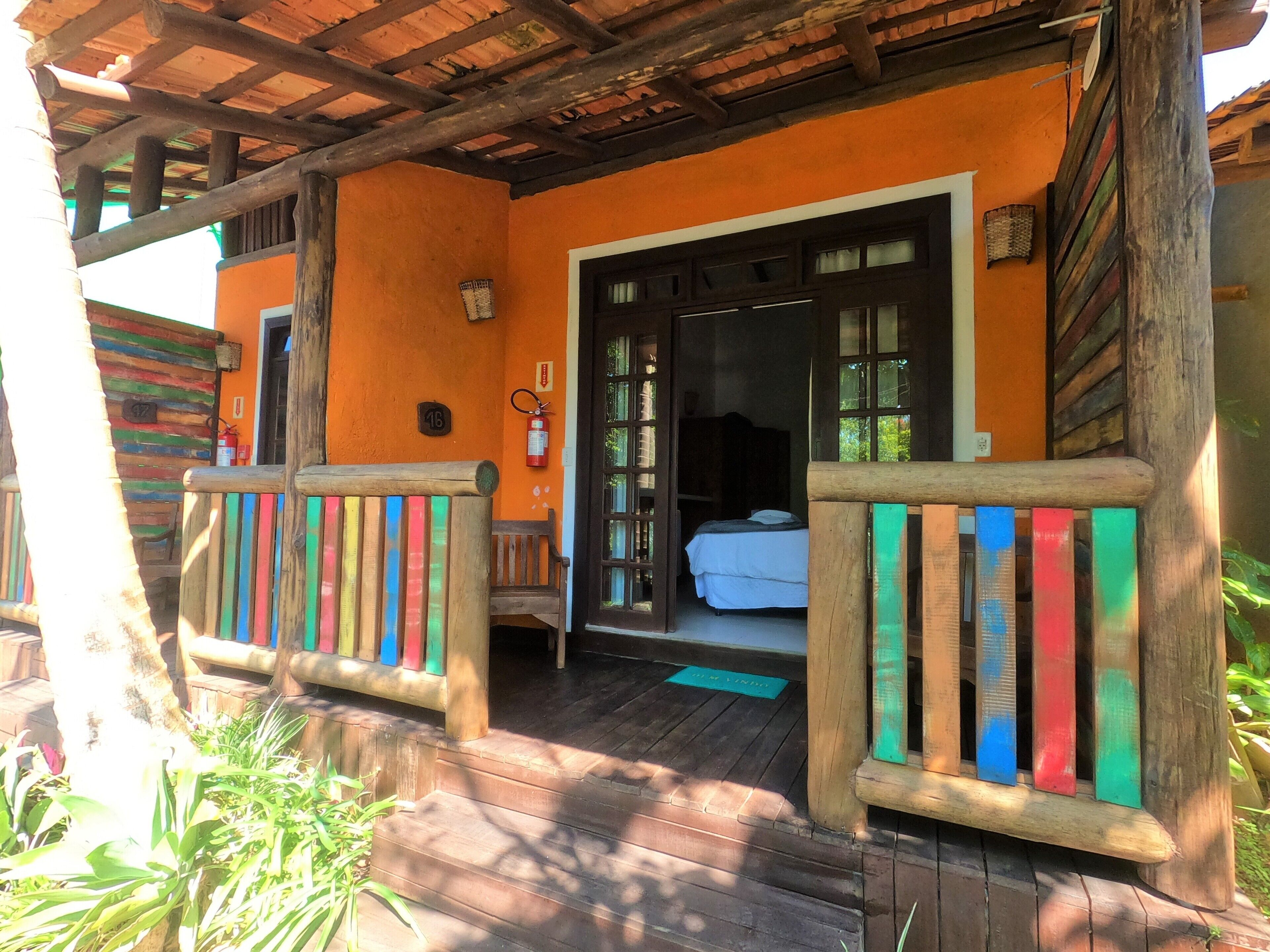 Une véranda en bois avec des balustrades colorées, une porte ouverte donnant sur une pièce avec un lit et une fenêtre, et un extincteur fixé au mur.