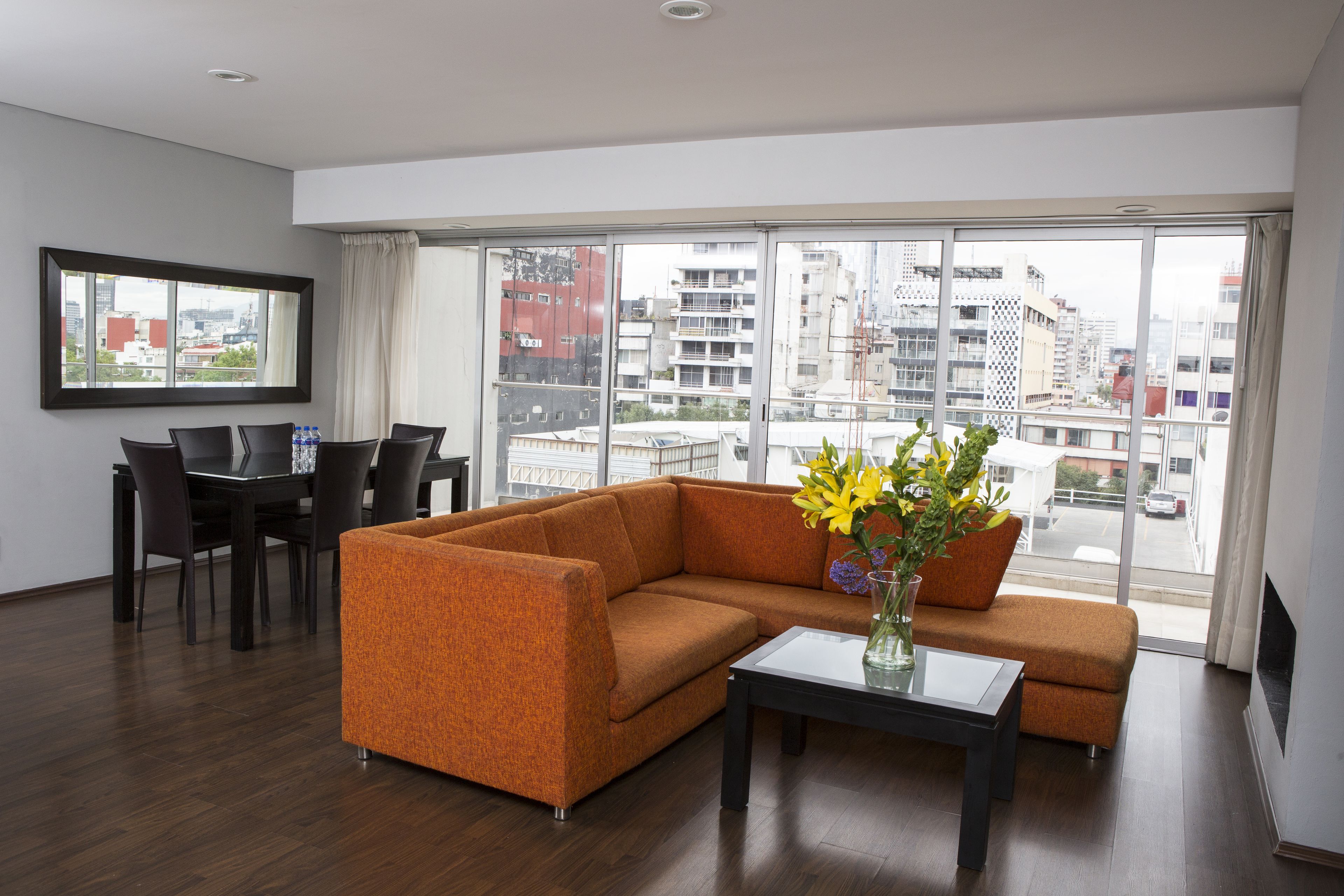 A modern living room with a sofa, a dining table, and chairs. There is a large window with a view of buildings.