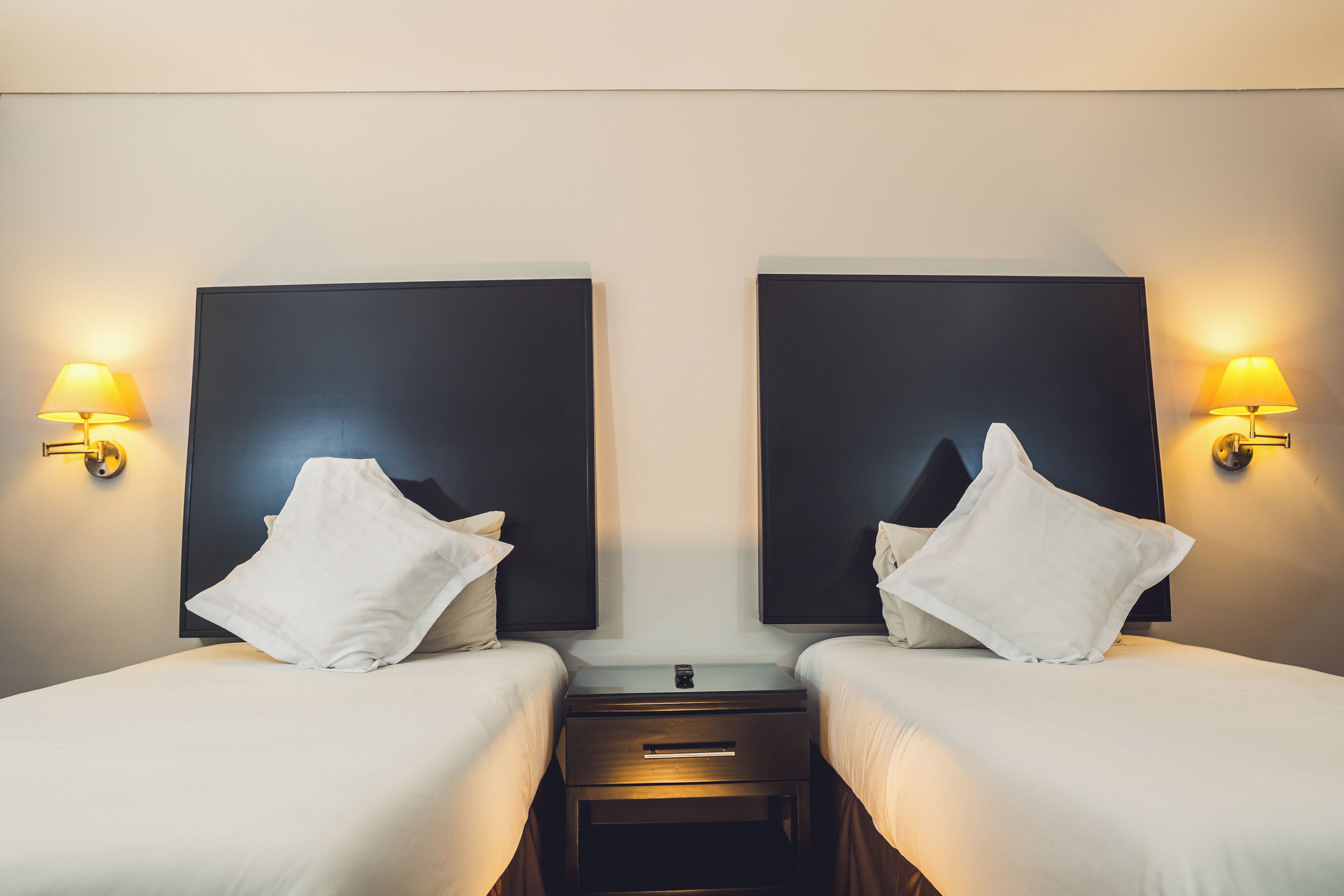 Habitación de hotel con dos camas, cada una con almohada blanca y una mesita de noche entre ellas.