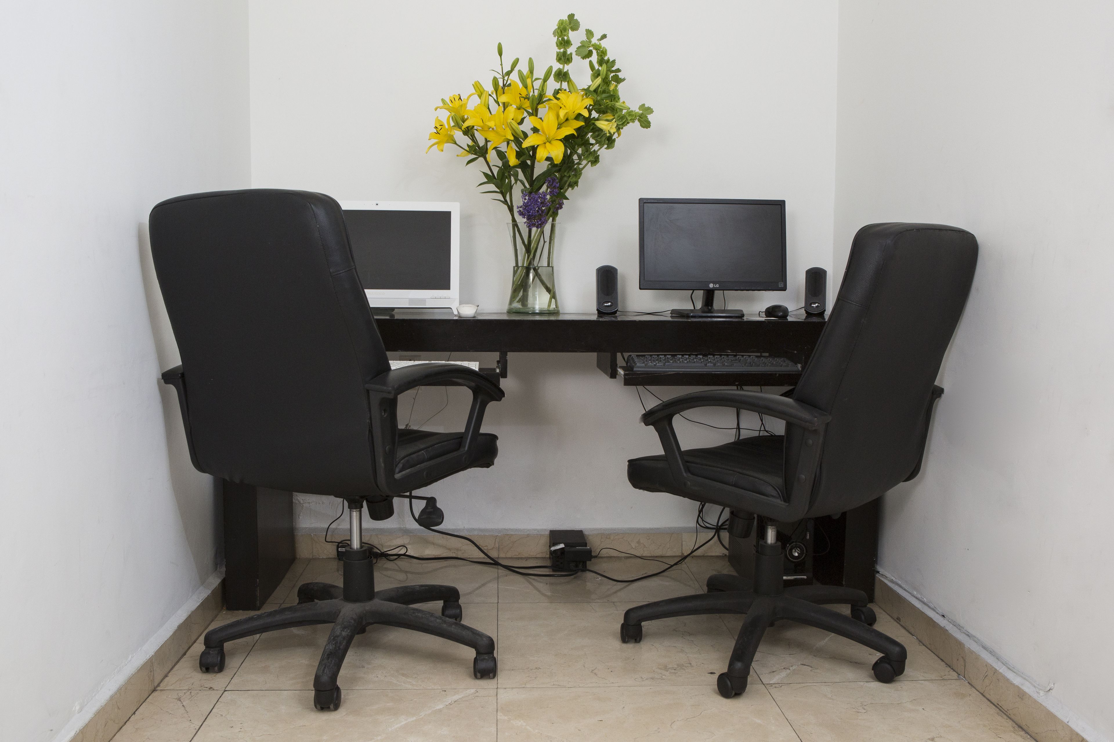A workspace with two black office chairs, a desk with two monitors, a keyboard, a mouse, and a vase with yellow flowers.