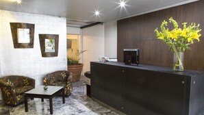 A reception area with a dark reception desk, two patterned armchairs, a small table with a vase of yellow flowers, and a potted plant. - Suites Ganges (Mexico City)