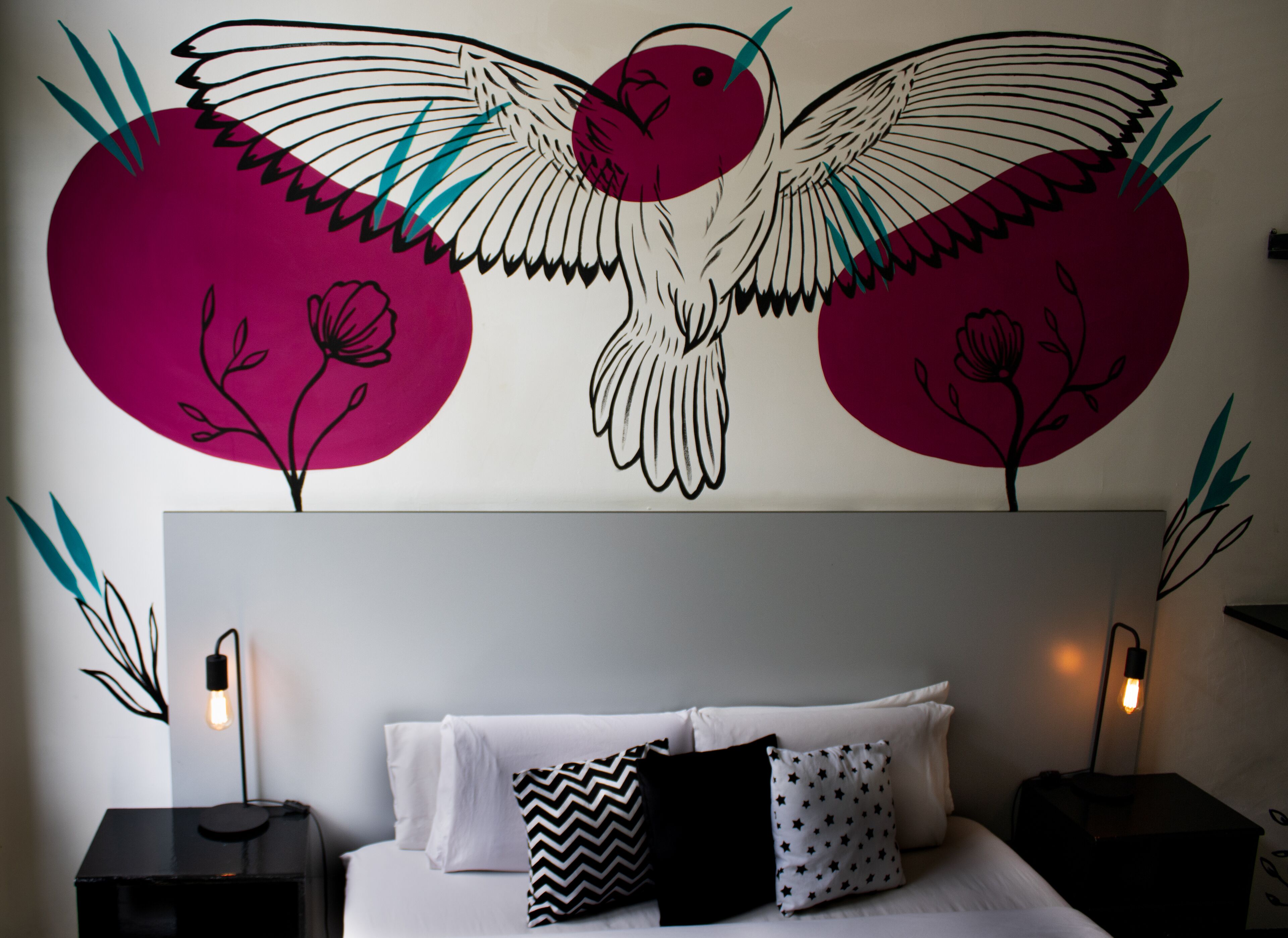 Un dormitorio con un gran mural de un pájaro, una cama con ropa de cama blanca y almohadas estampadas, y lámparas de noche.