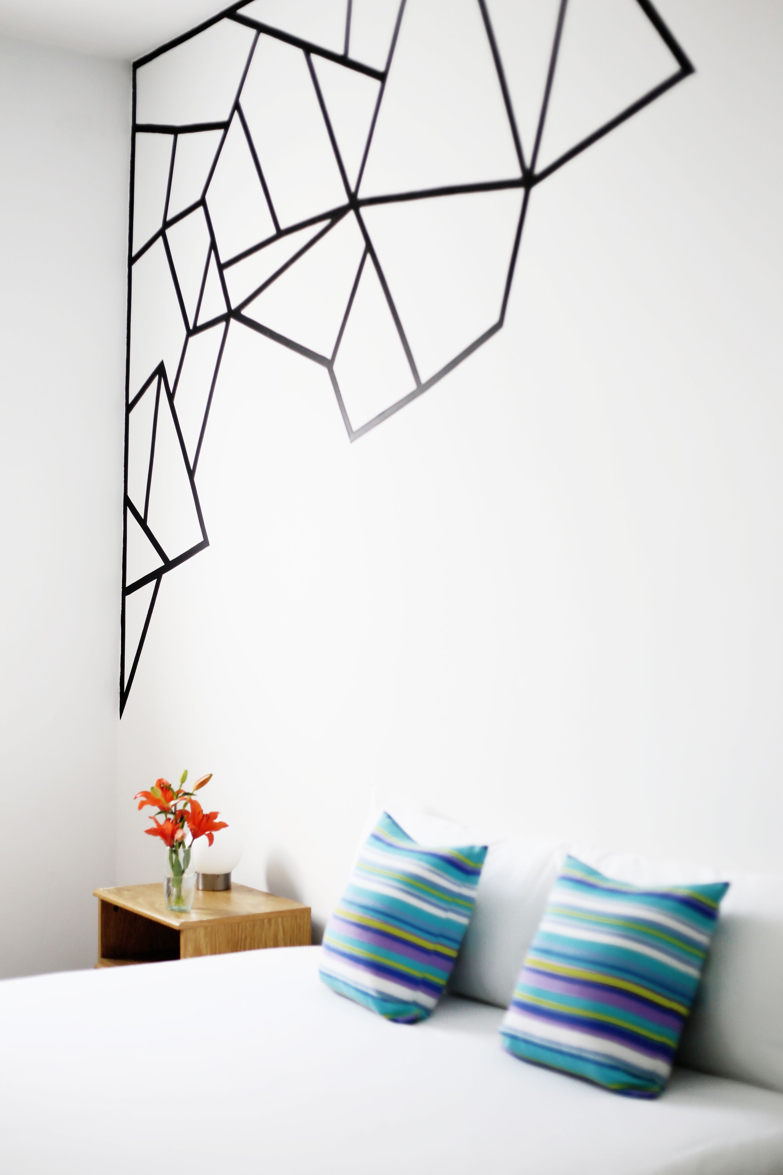 Un dormitorio moderno con una obra de arte geométrica en la pared, una mesita de noche con un jarrón de flores y almohadas a rayas sobre una cama blanca.