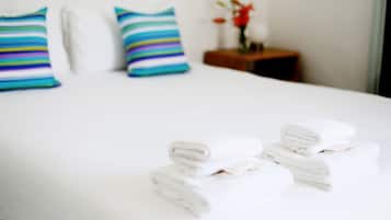Cama bem arrumada com lençóis brancos e almofadas listradas, uma mesinha de cabeceira com um vaso de flores e uma parede branca.
