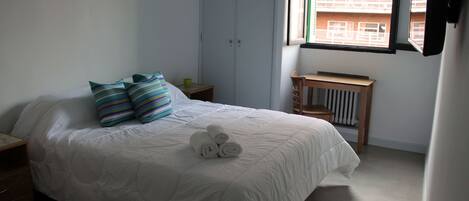 A bedroom with a bed, pillows, a bedside table, a radiator, and a window with a view of buildings.