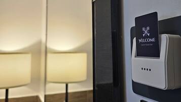 Camera d'albergo con un letto, un comodino, una lampada e una stazione di ricarica a muro con un cartello 'WELCOME'.