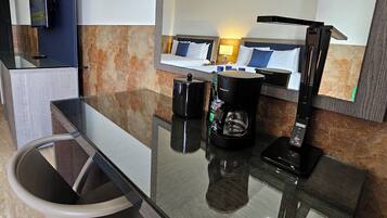 Ein modernes Hotelzimmer mit Glastisch, einem Sessel, einer Kaffeemaschine, einem am Wand montierten Flachbildfernseher und einem großen Spiegel, der den Raum widerspiegelt.
