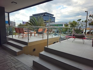 A modern outdoor seating area with steps leading up to a balcony, overlooking a cityscape with buildings and a bridge.