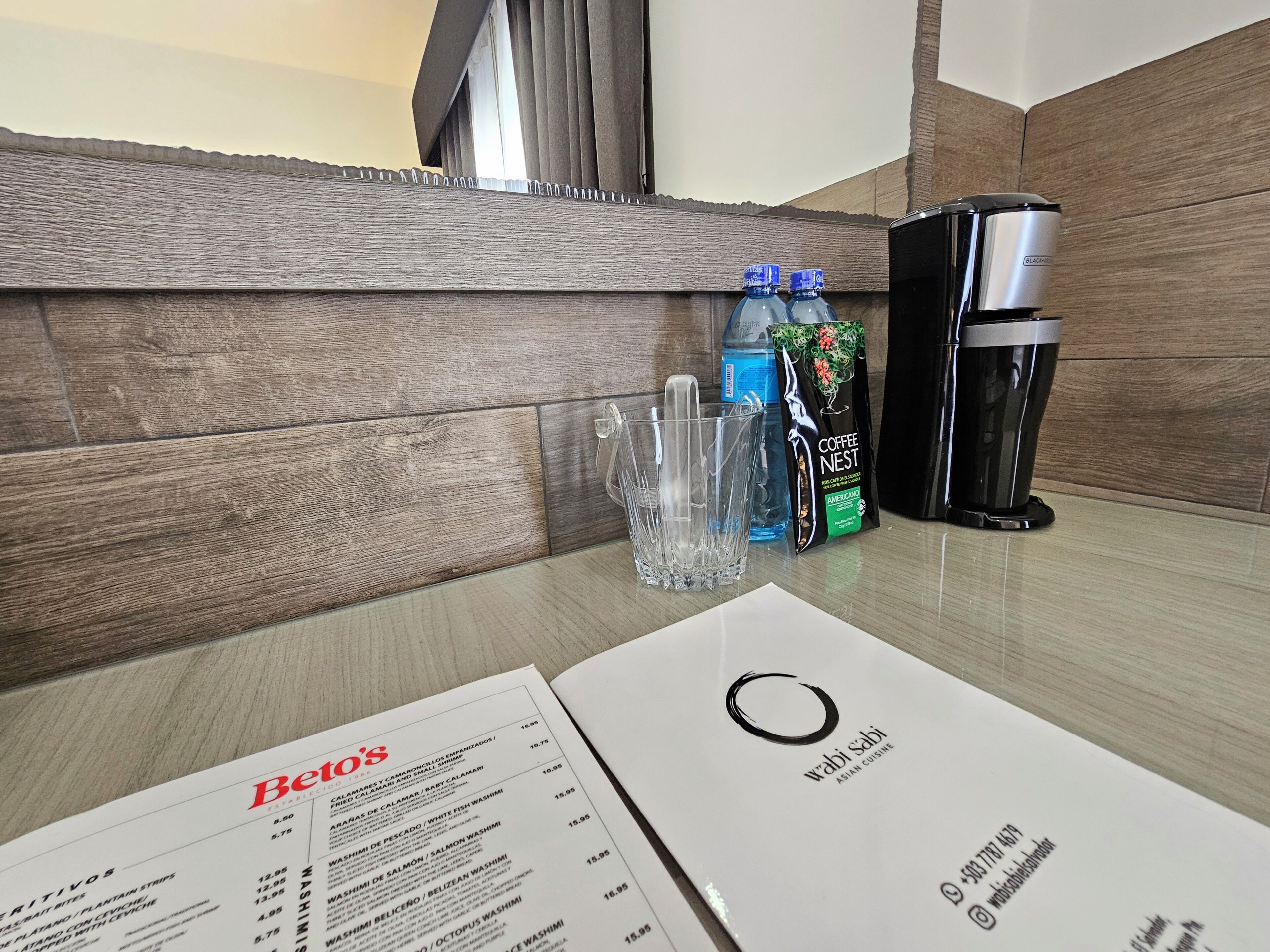 Een hotelkamer met een houten wand, een koffiezetapparaat, twee waterflessen, een glas en een menukaart.