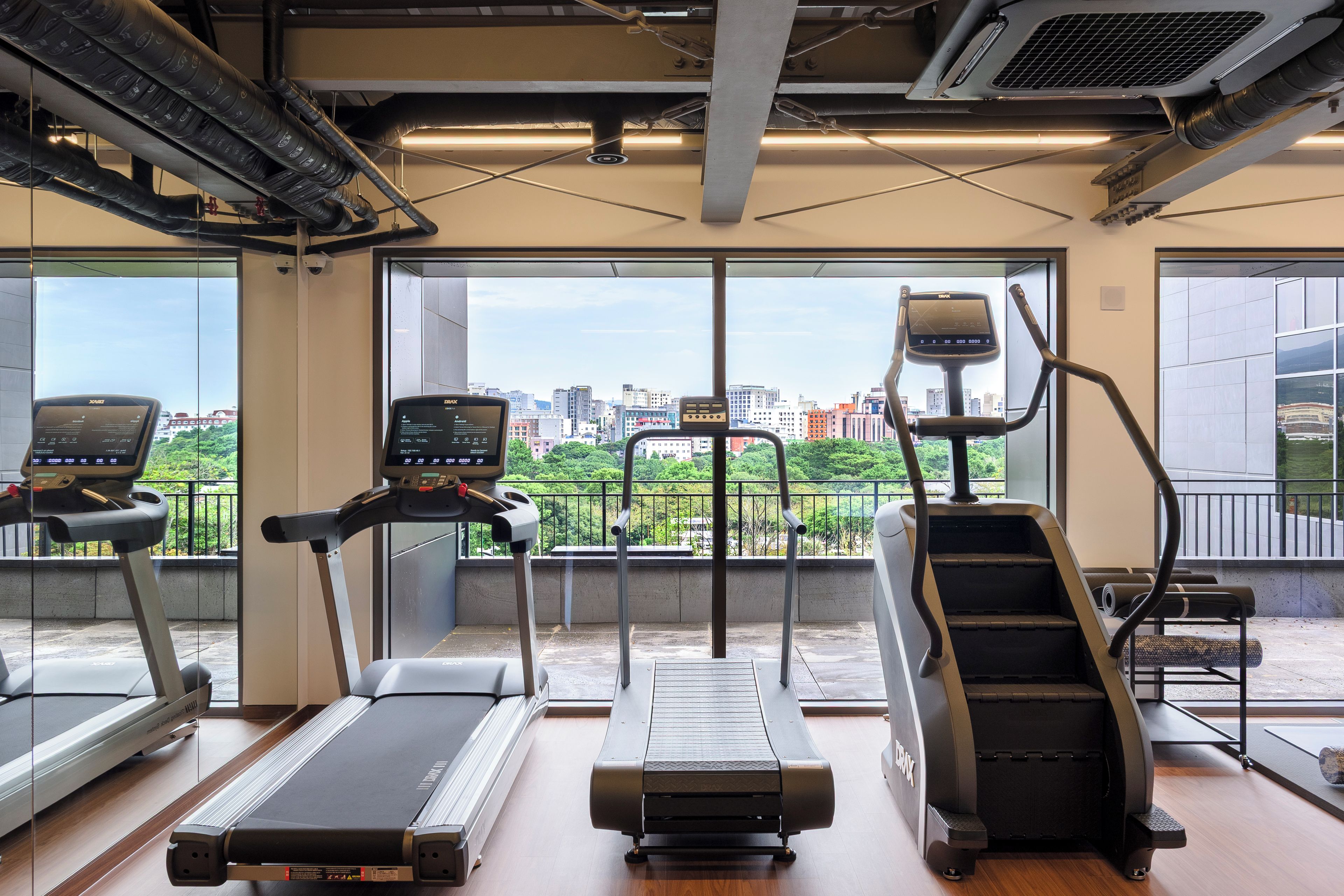 Ein Fitnessstudio mit Laufbändern, einem Steiger und einem Ergometer, mit Blick auf die Stadt durch große Fenster.